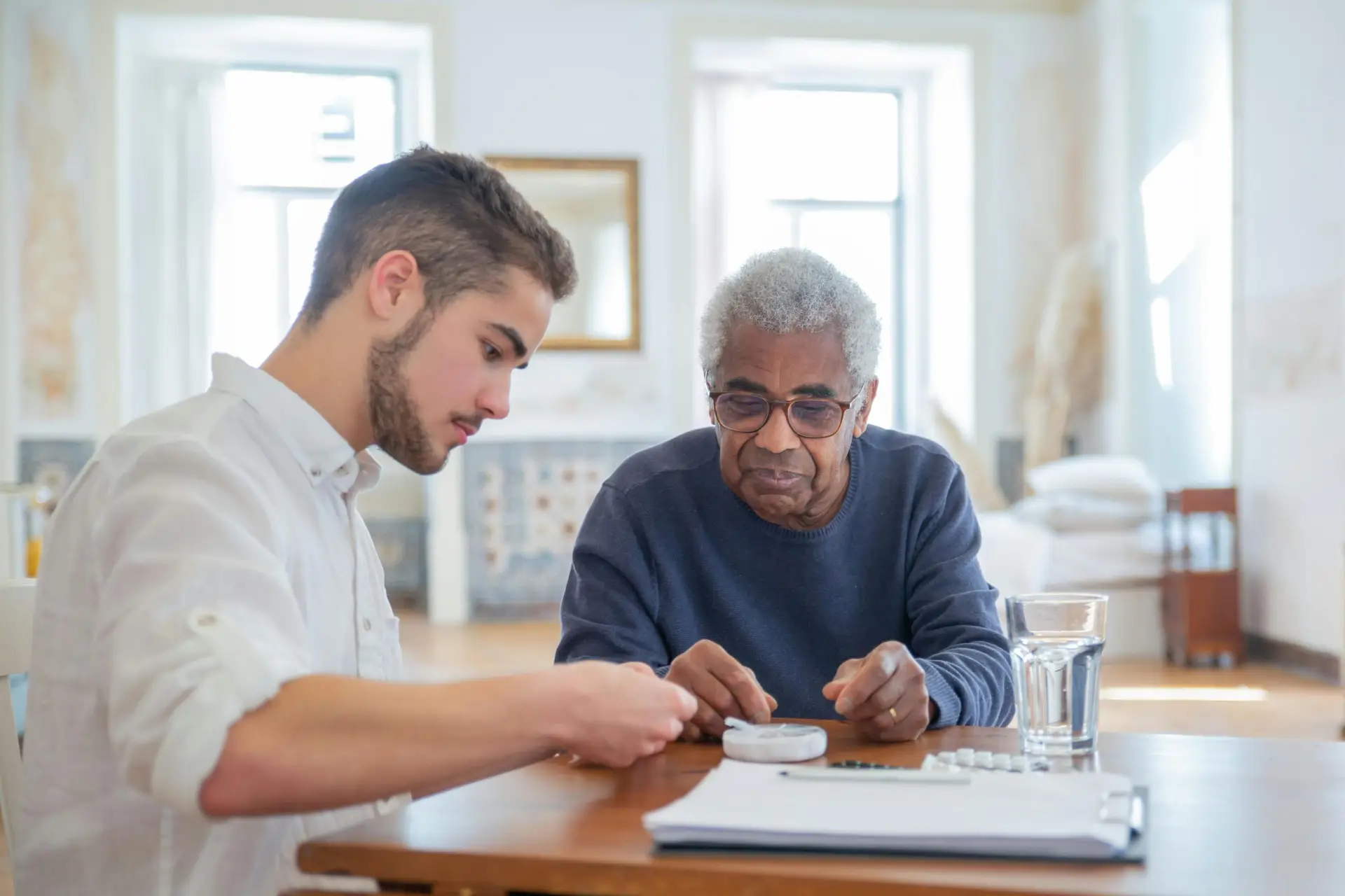 Young caregiver helping an elderly man organize and review his medications at a table with a pill organizer, notebook, and glass of water in a bright home setting.